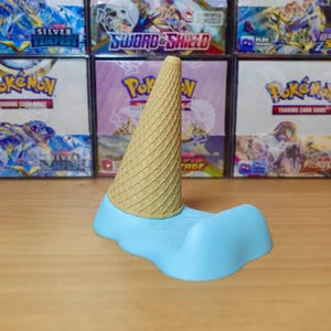 May include: A whimsical ice cream cone-shaped phone stand. The stand features a light blue base with a wavy design, supporting a tan, textured cone. The background includes various Pok&eacute;mon trading card game boxes.