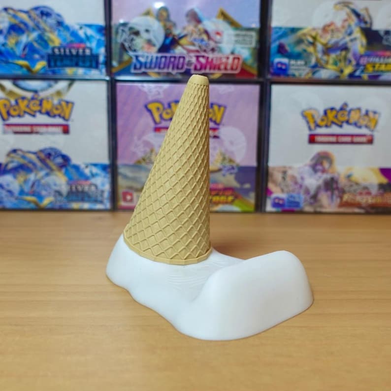 May include: A whimsical, white and tan ice cream cone-shaped phone holder. The holder features a textured, tan-colored cone atop a white, curved base, designed to cradle a phone. The background shows various Pokemon card boxes.