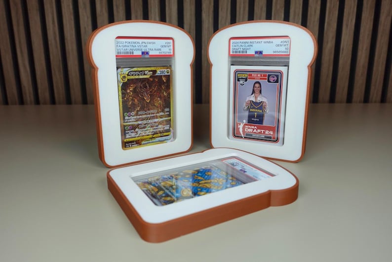 May include: Three rectangular display frames shaped like slices of bread with brown crust-colored edges and white centers. Each frame holds a PSA-graded trading card, including a gold Giratina VSTAR card and a Caitlin Clark WNBA Draft 24 card.