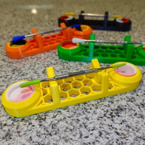 May include: A selection of brightly coloured dab tool holders, 3D-printed in yellow, orange, green, and black. Each holder has a different coloured tool. The holders feature a honeycomb pattern and two circular wells.