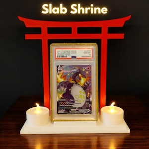 Może przedstawiać: Biały i czerwony ołtarz z napisem "Slab Shrine" nad ocenioną kartą Pokémon. Dwie świece na baterie flankują kartę. Karta przedstawia Charizard VMAX z zestawu Shining Fates.