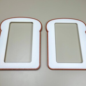 May include: Two flat, rectangular frames shaped like slices of bread. Each frame features a white center cutout surrounded by a brown border, mimicking the appearance of a crust. The objects are positioned side-by-side, displaying a consistent design.