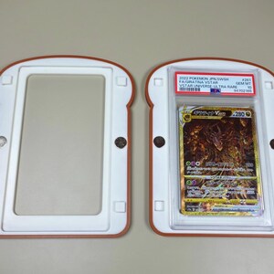 May include: A bread-shaped magnetic card display case with a white interior and brown border, shown open in two pieces. One side holds a 2022 Pokemon JPN SWSH #261 Giratina VSTAR VSTAR Universe Ultra Rare card graded Gem Mt 10 by PSA in a plastic slab.