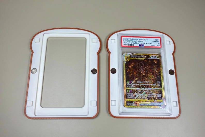 May include: A bread-shaped magnetic card display case with a white interior and brown border, shown open in two pieces. One side holds a 2022 Pokemon JPN SWSH #261 Giratina VSTAR VSTAR Universe Ultra Rare card graded Gem Mt 10 by PSA in a plastic slab.