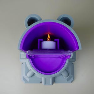 Pode incluir: Um casti&ccedil;al de parede cinza e roxo com uma vela branca acesa no interior. O suporte tem um topo arredondado com dois detalhes circulares e uma base retangular. O interior &eacute; de um roxo vibrante, criando um contraste marcante.