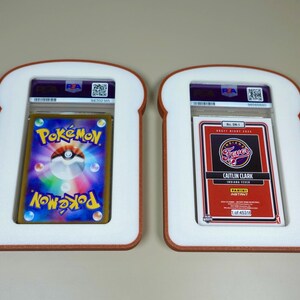 May include: Two PSA-graded trading cards encased in white, bread-shaped frames with brown crust-colored edges. The left card features a colorful Pok&eacute;mon logo and Pok&eacute; Ball graphic. The right card is a red Panini Instant Caitlin Clark Indiana Fever card.