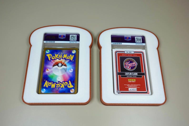 May include: Two PSA-graded trading cards encased in white, bread-shaped frames with brown crust-colored edges. The left card features a colorful Pok&eacute;mon logo and Pok&eacute; Ball graphic. The right card is a red Panini Instant Caitlin Clark Indiana Fever card.