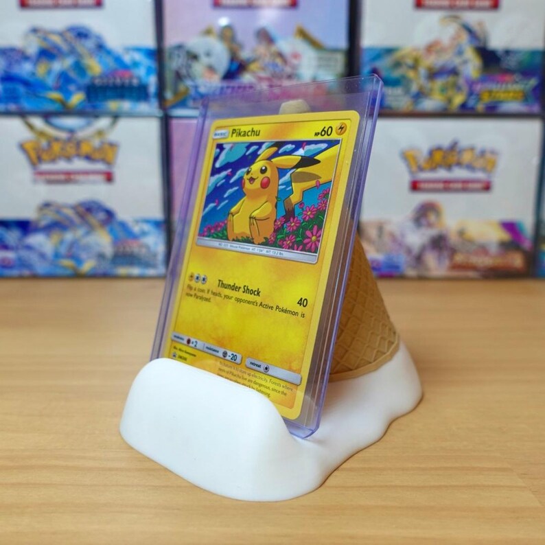 May include: A yellow Pikachu Pok&eacute;mon card in a protective sleeve, displayed on a white and tan ice cream cone-shaped stand. The card features a cartoon Pikachu character and the text "Pikachu".