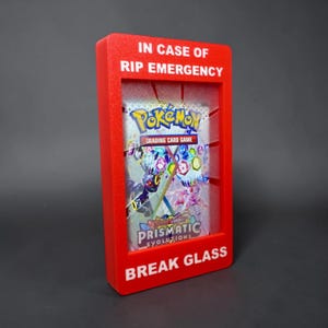 Peut inclure: Un boîtier de présentation rectangulaire rouge avec une façade transparente contenant un paquet de cartes à collectionner Pokemon Prismatic Evolutions. Le cadre porte le texte blanc IN CASE OF RIP EMERGENCY en haut et BREAK GLASS en bas.