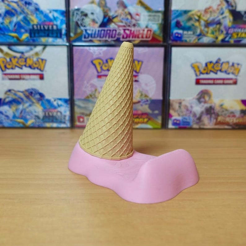 May include: A whimsical, pink ice cream-shaped phone stand with a waffle cone on top. The stand is designed to resemble melting ice cream. The background features boxes with colorful graphics.