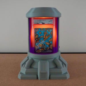 Pode incluir: Um expositor cinza e roxo com um interior laranja brilhante, exibindo um cart&atilde;o colecion&aacute;vel Pok&eacute;mon. O cart&atilde;o est&aacute; em uma capa protetora, e o expositor tem um design futurista com formato cil&iacute;ndrico e uma base.