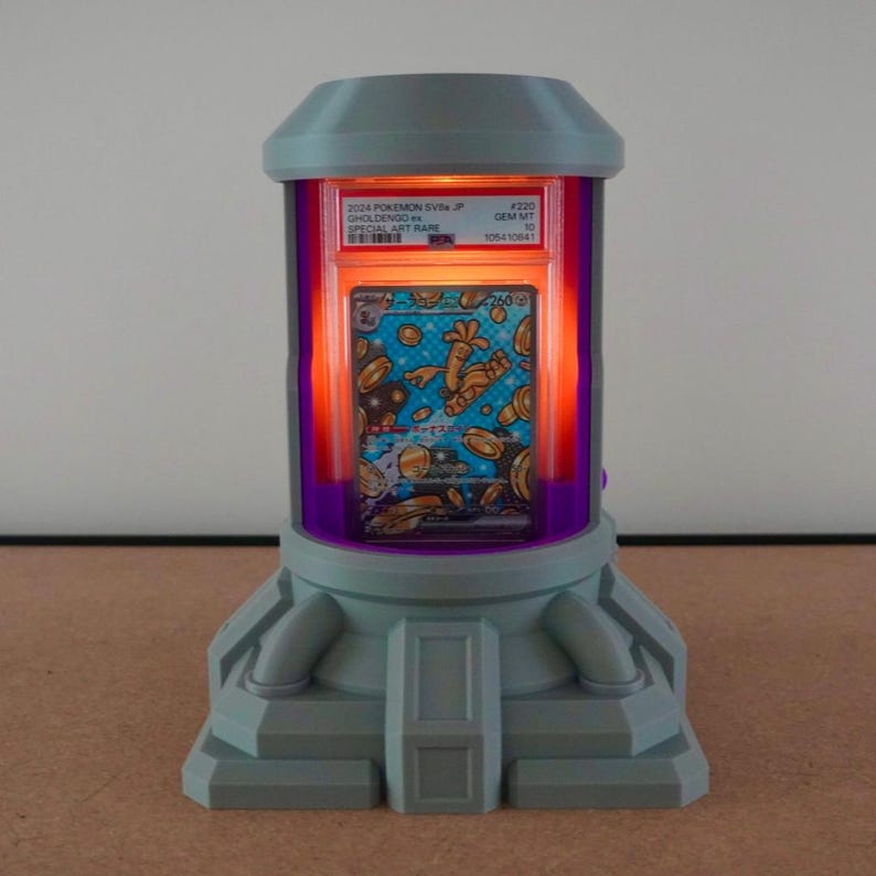 Pode incluir: Um expositor cinza e roxo com um interior laranja brilhante, exibindo um cart&atilde;o colecion&aacute;vel Pok&eacute;mon. O cart&atilde;o est&aacute; em uma capa protetora, e o expositor tem um design futurista com formato cil&iacute;ndrico e uma base.
