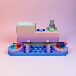 Puede incluir: Un organizador multicolor impreso en 3D con una base degradada rosa y morada. Cuenta con un portalápices, dos bandejas circulares con detalles de arcoíris y una pieza de vidrio transparente. La base es azul con un patrón de panal.