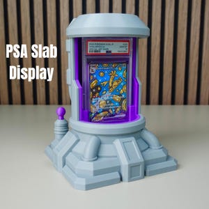 Pode incluir: Um expositor de placas PSA cinza com interior roxo, segurando um cart&atilde;o Pok&eacute;mon. O expositor tem um design futurista com o texto "PSA Slab Display" no lado esquerdo. O cart&atilde;o &eacute; vis&iacute;vel dentro.