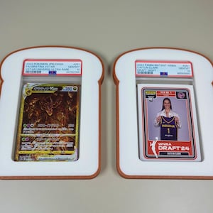May include: Two PSA graded trading cards encased in white, bread-shaped frames with brown crust-colored borders. The left card is a gold-toned 2022 Pokemon Giratina VSTAR, and the right card is a 2024 Panini Instant WNBA Caitlin Clark Draft Night card.