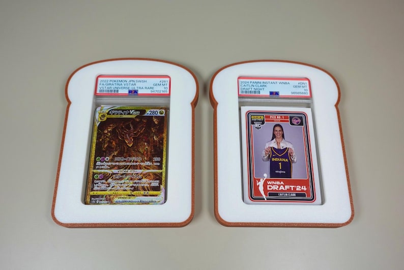 May include: Two PSA graded trading cards encased in white, bread-shaped frames with brown crust-colored borders. The left card is a gold-toned 2022 Pokemon Giratina VSTAR, and the right card is a 2024 Panini Instant WNBA Caitlin Clark Draft Night card.