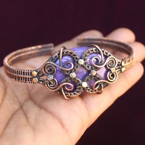 Pode incluir: Um bracelete envolto em fio de cobre com uma pedra roxa engastada no centro. A pedra é cercada por intrincados desenhos de fio e pequenos detalhes dourados.