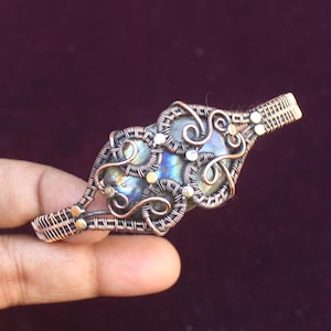 May include: A copper wire wrapped bracelet with a large iridescent labradorite gemstone. The wire is twisted and formed into a delicate design.