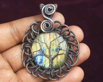 May include: A copper wire wrapped pendant featuring a blue and yellow labradorite gemstone with a tree design. The pendant is on a hand.