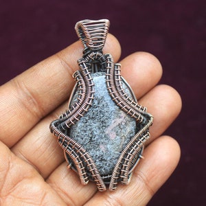 Peut inclure: Un pendentif en fil d'argent enroulé autour d'une pierre grise et rose. Le pendentif est de forme ovale et présente un délicat motif de fil.