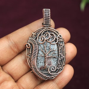 以下が含まれることがあります： 木製のデザインが施されたグレーとホワイトの石を、銅線で包んだペンダント。ペンダントは楕円形で、銅製のベイルが付いています。