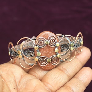 May include: A copper and brass wire wrapped bracelet with a large oval-shaped sandstone stone in the center. The stone is surrounded by wire spirals and small brass beads.