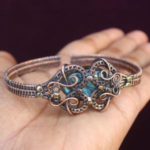 May include: A copper wire wrapped bracelet with a blue-green labradorite gemstone in the center. The bracelet is decorated with small gold beads.