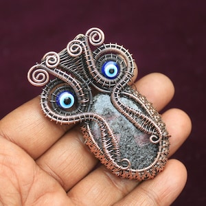 Może przedstawiać: Zawieszka owinięta miedzianym drutem z szarym i różowym kamieniem i dwoma niebieskimi talizmanami w kształcie oka.