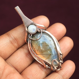 May include: A copper wire wrapped pendant featuring a large iridescent labradorite gemstone and a smaller white moonstone. The pendant has a delicate, intricate design.