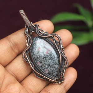 Könnte beinhalten: Ein Anhänger aus Kupferdraht, der mit einem grauen und rosafarbenen Edelstein umwickelt ist. Der Anhänger ist oval geformt und hat ein filigranes Drahtdesign.