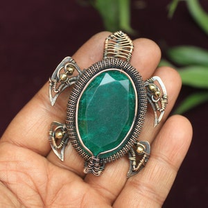 May include: A green gemstone turtle pendant made of wire wrapped around a green stone. The turtle is made of silver and gold wire.