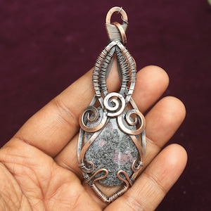 May include: A silver and copper wire wrapped pendant featuring a grey and pink speckled stone. The pendant is shaped like a teardrop with intricate wire designs.