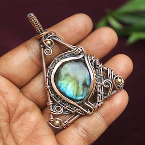 Könnte beinhalten: Ein Anhänger aus umwickeltem Kupferdraht mit einem blaugrünen Labradorit-Edelstein. Der Anhänger hat die Form eines Dreiecks und oben eine kleine goldene Perle.