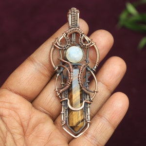 May include: A copper wire wrapped pendant featuring a tiger's eye stone and a moonstone. The pendant is in the shape of a geometric design with a copper bail.