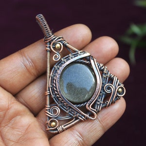 May include: A triangular wire-wrapped pendant with a gold-flecked green stone set in the center. The pendant is made of copper wire and has a delicate, intricate design.