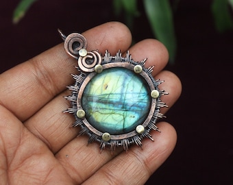 Puede incluir: Colgante artesanal con una piedra preciosa redonda e iridiscente en el centro. La piedra está rodeada por un marco de metal color cobre con puntas radiantes y pequeños detalles de latón. Tiene un lazo para una cadena.