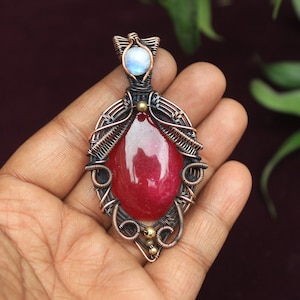 Puede incluir: Un colgante de piedra roja con un envoltorio de alambre de cobre y un pequeño acento de piedra lunar. El colgante se sostiene en una mano.
