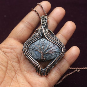 Op de afbeelding: Een hanger omwikkeld met koperdraad met een grijze en witte agaat-steen met een boomontwerp. De hanger hangt aan een koperen ketting.