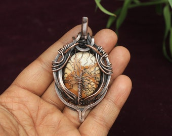May include: A copper wire wrapped pendant with a brown and white agate stone in the center. The pendant is in the shape of a tree and has a copper bail.