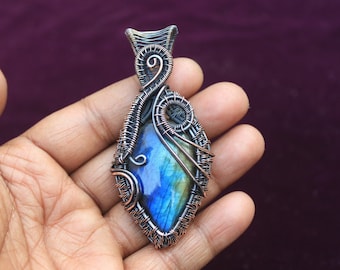 Puede incluir: Un colgante de piedra de labradorita azul y verde envuelto en alambre de plata con diseños intrincados.