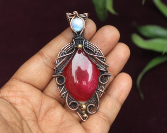 May include: A red gemstone pendant with a copper wire wrap and a small moonstone accent. The pendant is held in a hand.