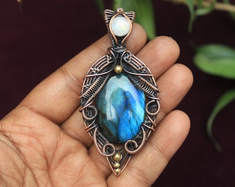 Puede incluir: Un colgante envuelto en alambre de cobre con una piedra preciosa de labradorita azul y un acento de ópalo blanco. El colgante se sostiene en una mano.