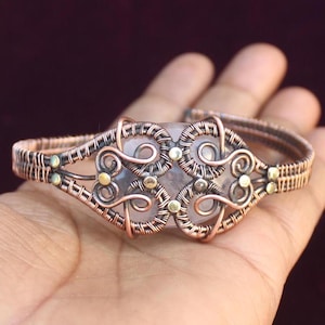 Könnte beinhalten: Ein Armband aus umwickeltem Kupferdraht mit einem klaren Quarzstein in der Mitte. Das Armband hat ein filigranes, kompliziertes Design mit kleinen goldenen Akzenten.