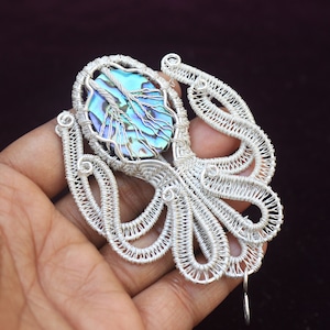 May include: A silver wire pendant with an abalone shell centre, displaying iridescent blues and greens. The wire is crafted into an octopus shape. The pendant is held in a hand against a dark background. The pendant's design is intricate.