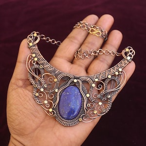 Op de afbeelding: Een handgemaakte ketting met een grote, ovale, diepblauwe edelsteen in het midden. Het ingewikkelde ontwerp bevat koperkleurig draad met decoratieve accenten en een kettingsluiting. De ketting wordt op een hand getoond.