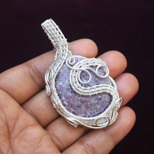 May include: A silver wire wrapped pendant featuring a light purple and gray gemstone. The pendant is intricately designed with swirls and loops of wire.