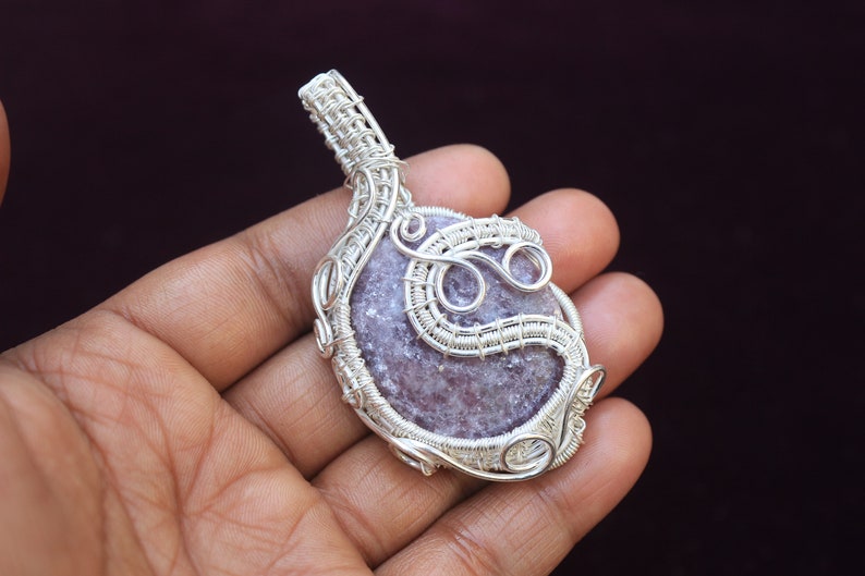 May include: A silver wire wrapped pendant featuring a light purple and gray gemstone. The pendant is intricately designed with swirls and loops of wire.