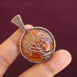 Peut inclure: Pendentif rond avec un motif d'arbre. Il présente un cadre en fil de couleur cuivre et une pierre centrale de couleur ambre. Le motif de l'arbre est réalisé en fil fin, et le pendentif est suspendu par une boucle en fil.