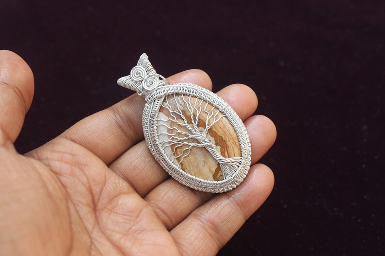 May include: Silver wire wrapped pendant featuring a brown and white tree of life stone. The pendant is oval shaped and has a silver bail.