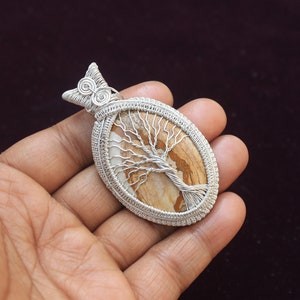 May include: Silver wire wrapped pendant featuring a brown and white tree of life stone. The pendant is oval shaped and has a silver bail.